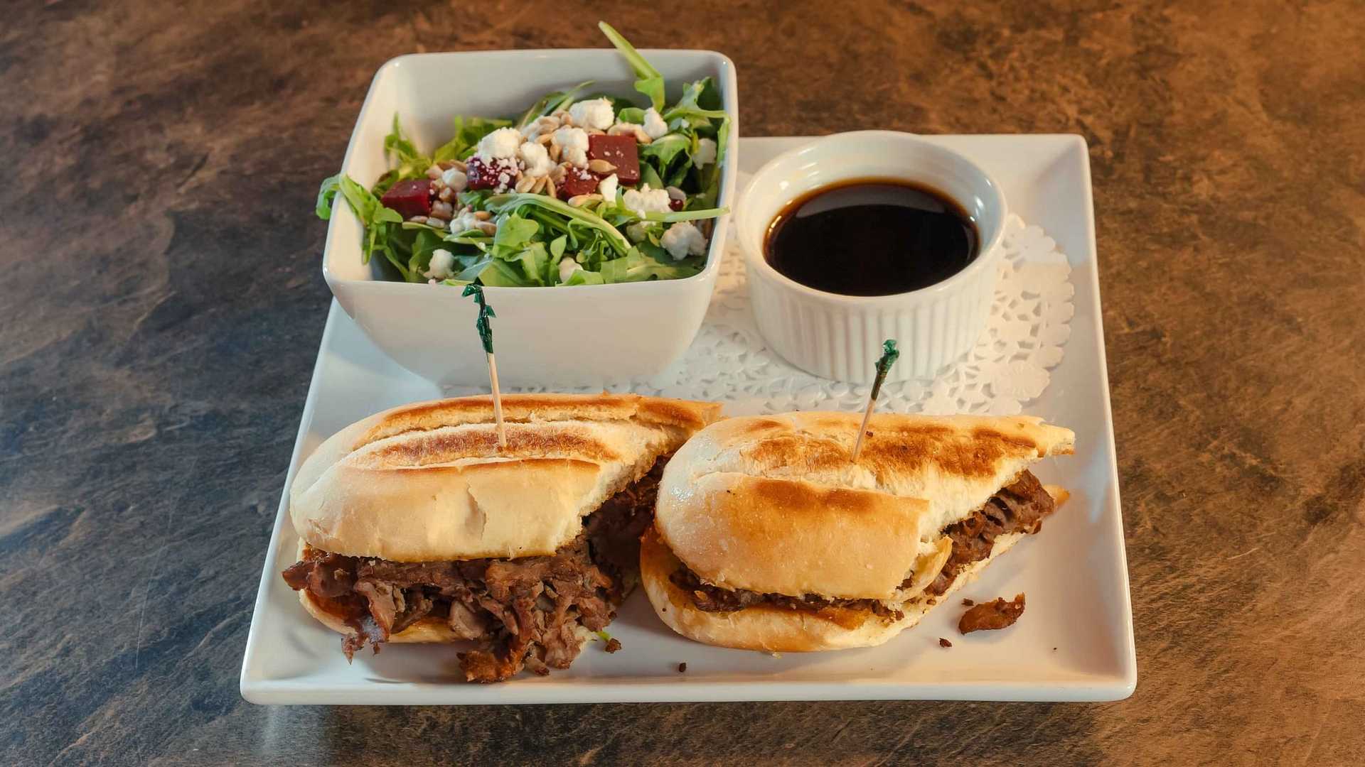 Beef sandwiches with a side of fresh salad and dipping sauce on a square plate.