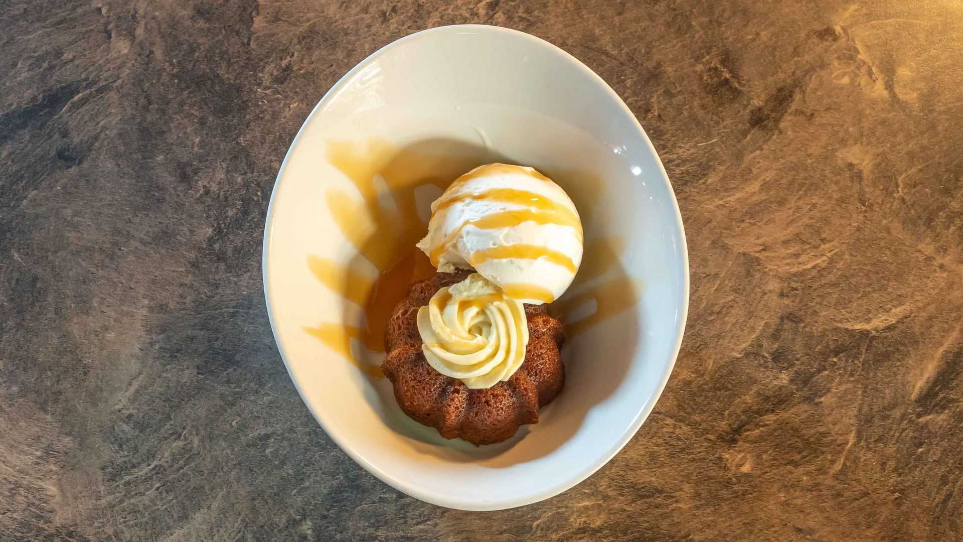 Dessert plate with a caramel-drizzled ice cream scoop and a small Bundt cake on a textured surface.