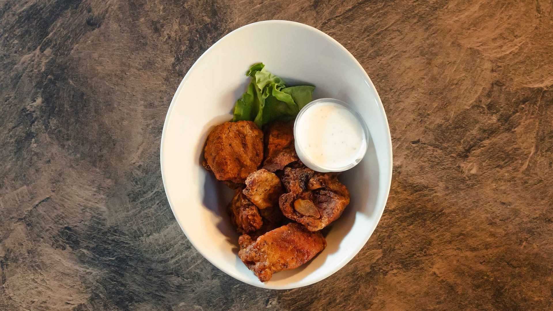 White bowl of crispy fried chicken pieces, greens, and a cup of dipping sauce on a textured surface.