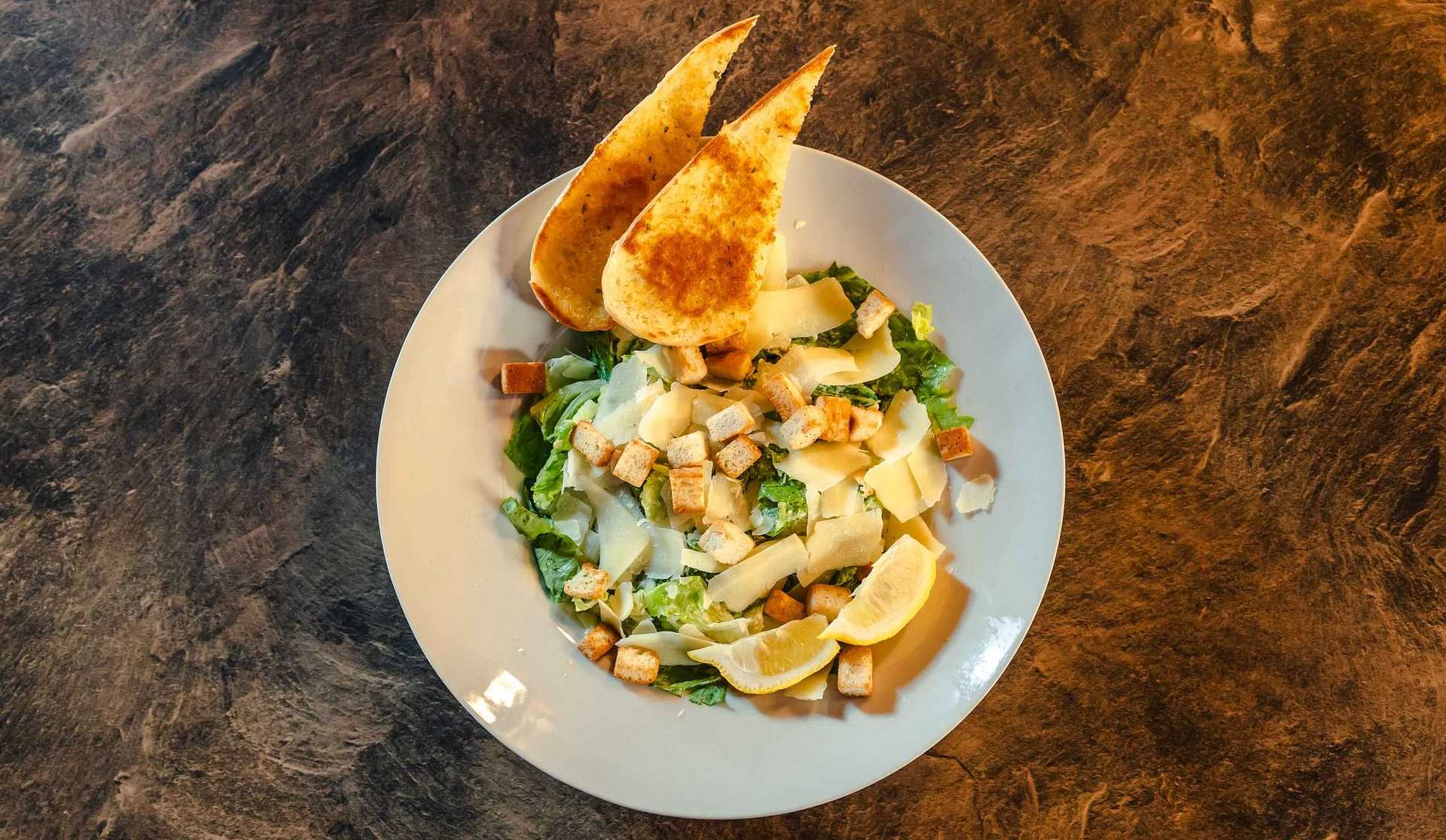 Caesar salad with croutons, parmesan, lemon wedges, and toasted bread on a textured surface.